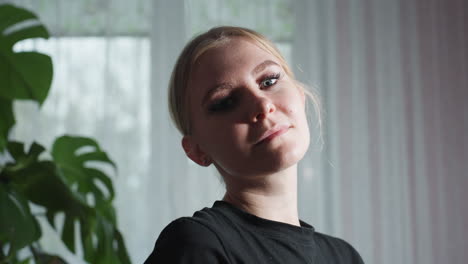 close up of massage therapist in black shirt gazing at camera with serious expression, standing beside large leafy indoor plant, with soft daylight filtering through sheer curtains in background