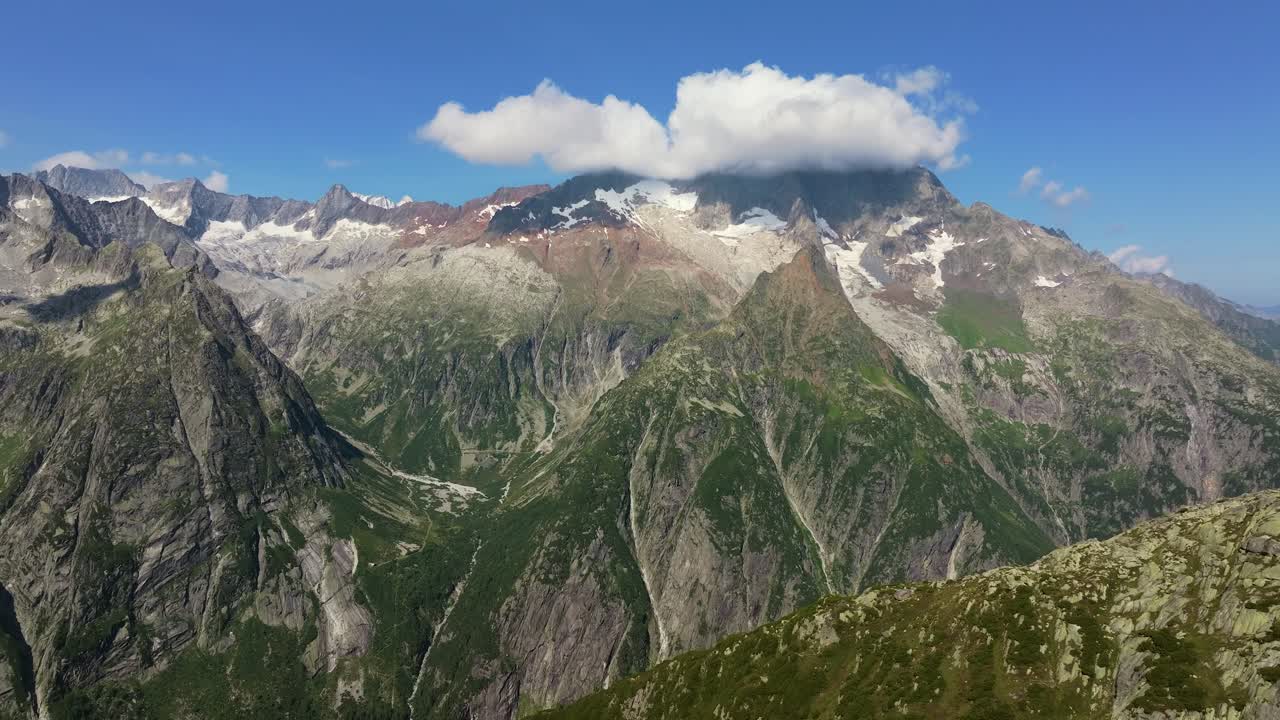 Drone shot of majestic alpine peaks with steep rocky cliffs, green valleys, and snow patches under a clear blue sky. A large cloud hovers above the rugged mountain summits