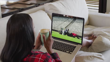compuesto de una mujer sentada en casa sosteniendo un café viendo un partido de rugby en una computadora portátil