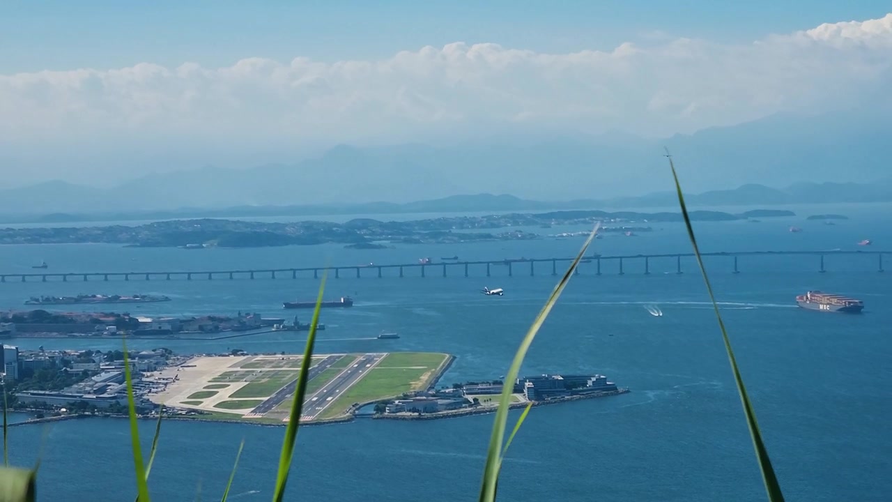 Vista panoramica dal Pan di Zucchero sull'aeroporto Santos Dumont e sul Centro di Rio de Janeiro, con il Ponte Rio–Niterói che si estende sulla Baia di Guanabara e un aereo che decolla in una giornata di sole.