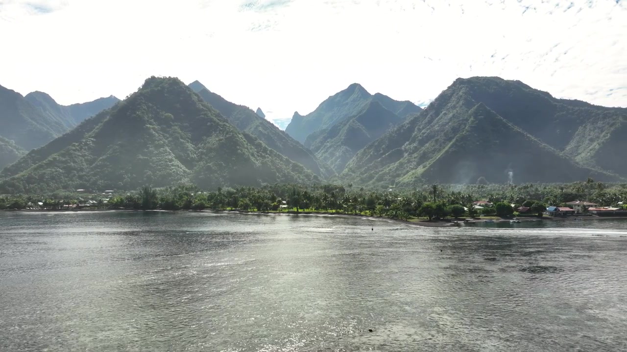 Jagged peaks rise above lush slopes and shoreline village at Teahupo'o, Tahiti, French Polynesia, calm lagoon stretches beneath misty morning light and drifting clouds. Aerial drone view.