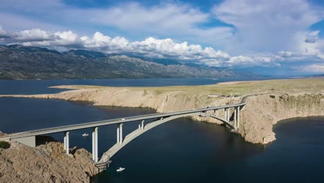 Paski-Most---Barco-Navegando-Bajo-El-Puente-Pag-Con-Vistas-Panorámicas-A-La-Montaña-Bajo-Nubes-Blancas-En-Croacia