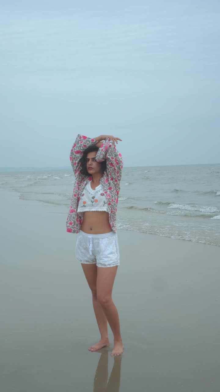 Young woman confidently standing on a wet sandy beach, hands on hips, wearing a patterned top and shorts, enjoying ocean waves and a cloudy sky during summer vacation