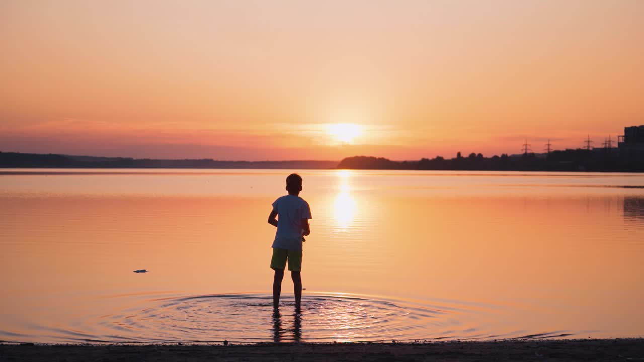 Backside view of a boy in the evening. Child standing in water and launching paper plane into the river at sunset. Slow motion.