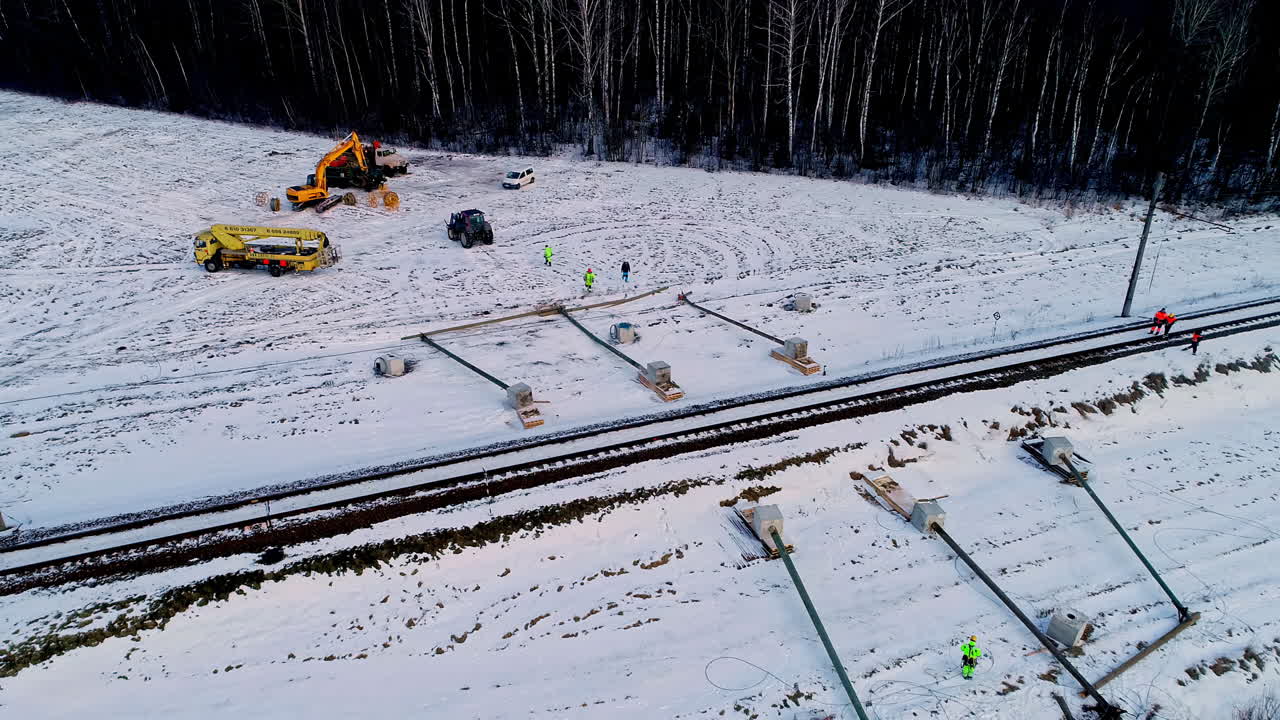 Men And Equipment At Work At The Installation Of Utility Poles And Electric Cables Along Railway On A Snowy Landscape In Winter