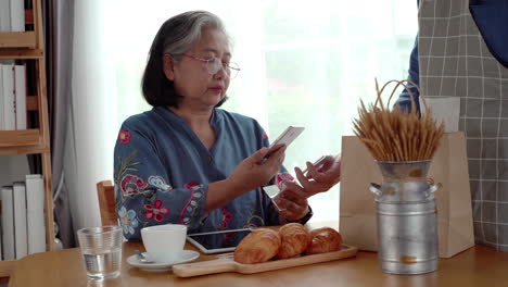 Active-senior-Asian-woman-working-in-a-coffeeshop-using-a-mobile-device,-smartwatch-for-NFC,-QR-code-scanning,-contactless-payment-to-pay-for-coffee-and-snack