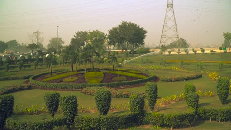 El-Aspersor-De-Impulso-Rociando-El-Jardín,-Hermoso-Diseño-Del-Jardín-Con-Varias-Plantas-Y-árboles,-Caminos-En-La-Parte-Trasera-Del-Parque,-El-Tráfico-Continúa