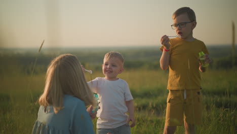 a woman in a blue dress kneels in a grassy field at sunset, holding a bubble bottle while her younger son blows bubbles from the wand. her older son, dressed in a yellow outfit, also blows bubbles