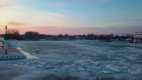 Una-Toma-Panorámica-Alrededor-De-Un-Faro-Y-Un-Muelle-Revela-Una-Hermosa-Puesta-De-Sol-Roja-Y-Naranja-Sobre-El-Lago-Congelado-En-Canadá