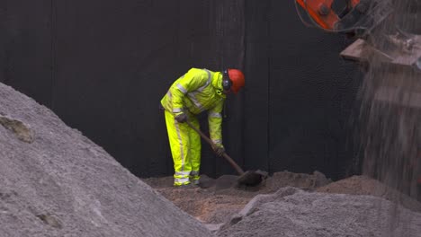 Arbeiter-Gräbt-Mit-Schaufel,-Während-Im-Vordergrund-Der-Bagger-Jede-Menge-Sand-Schüttet