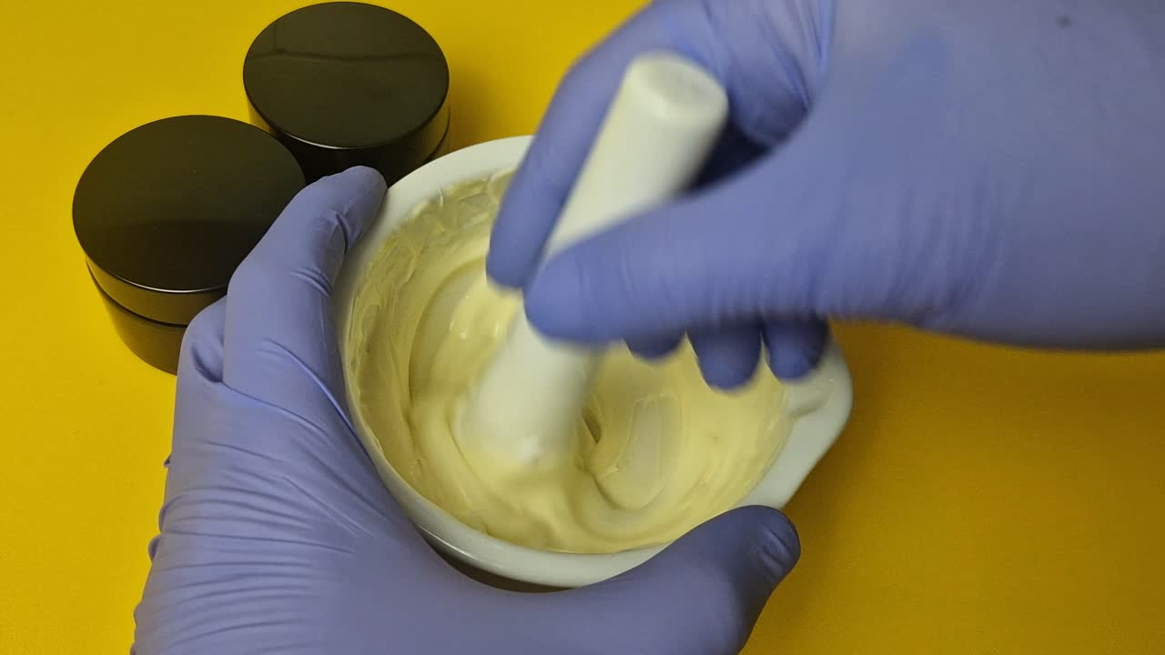 Extreme close-up view of a cosmetic chemist mixing a luxurious, yellow cream base in a white ceramic mortar with a pestle. Hands wear protective blue gloves. Perfect for beauty, pharmacy, and skincare