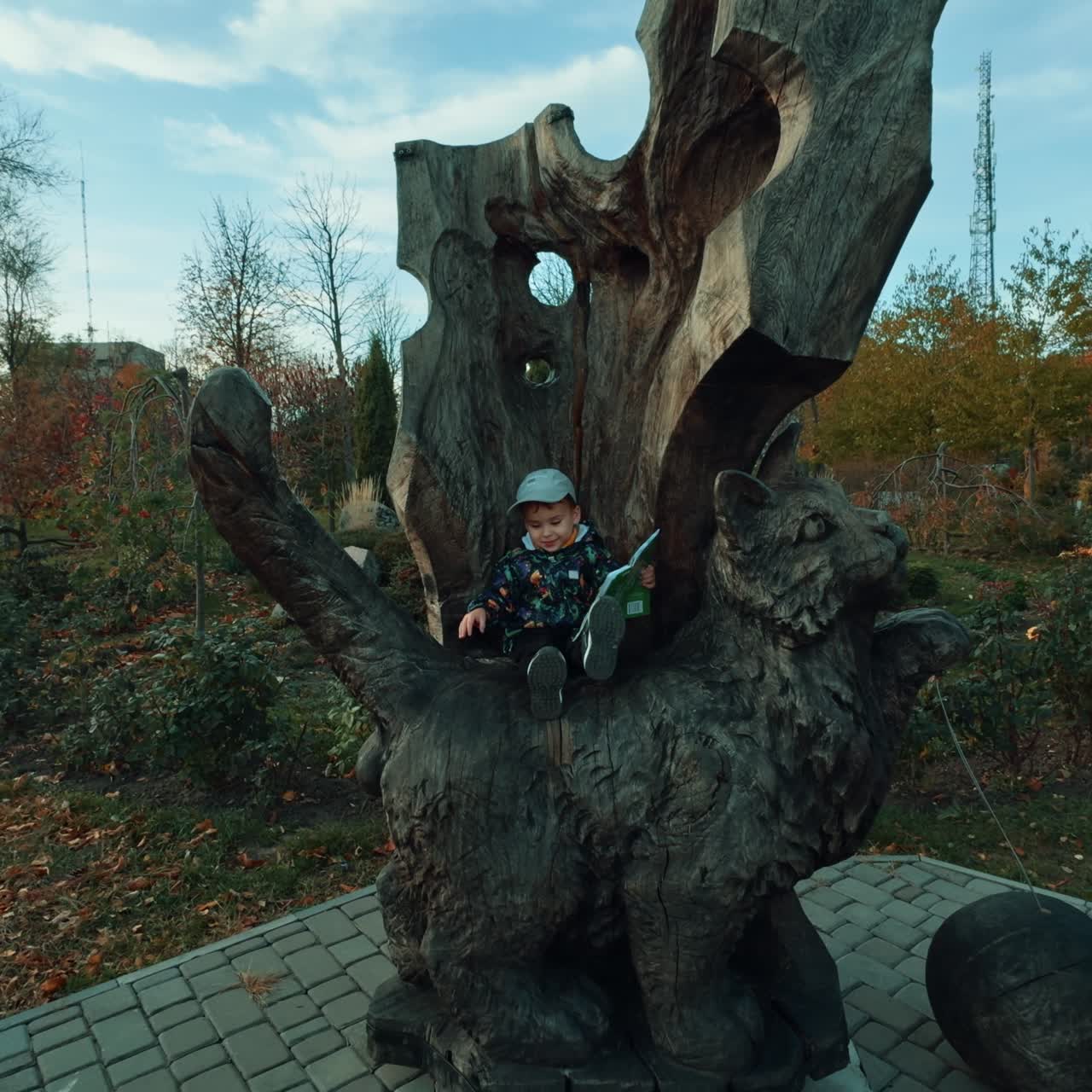 Gutt som sitter på trestolen som er skåret ut fra et tre. Nydelig baby holder en bok. Høstpark i bakgrunnen