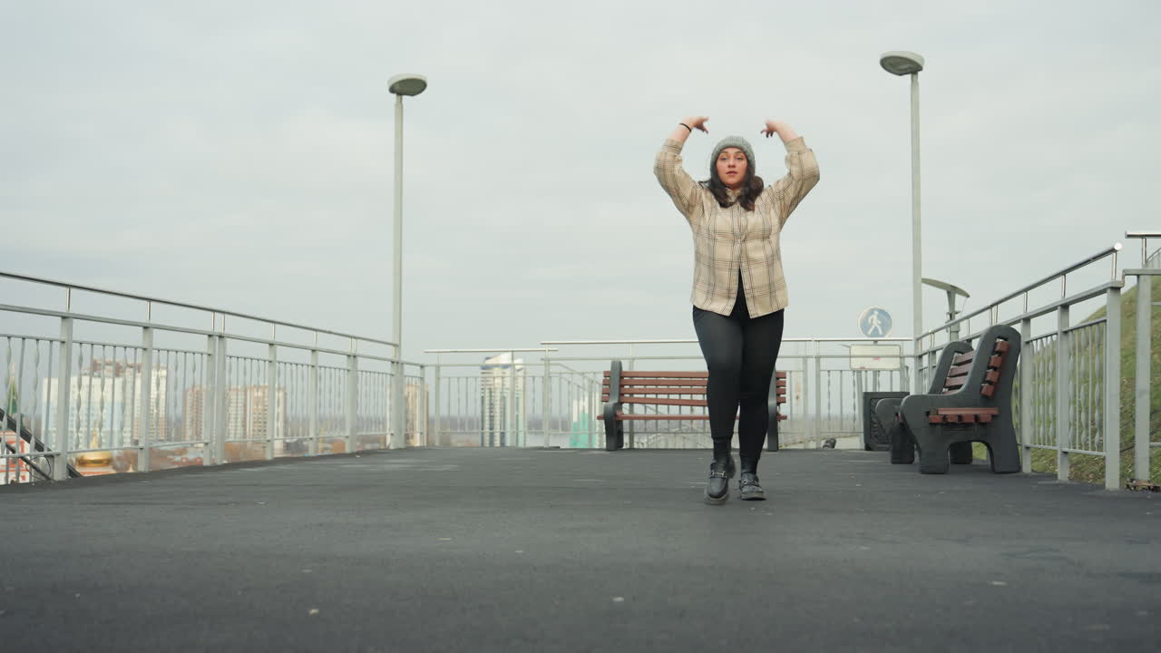 Mulher no telhado dançando sob céu nublado, casaco de inverno e gorro de lã, performance de rua expressiva perto de banco e corrimão, paisagem urbana ao fundo, giros e poses enérgicos, movimento alegre e espontâneo, cativante.