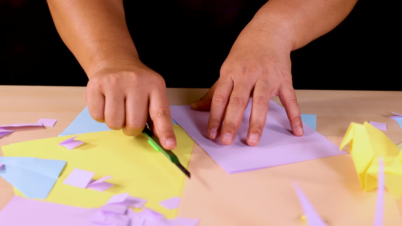 Person uses craft knife to cut pastel paper on desk, surrounded by origami and scraps