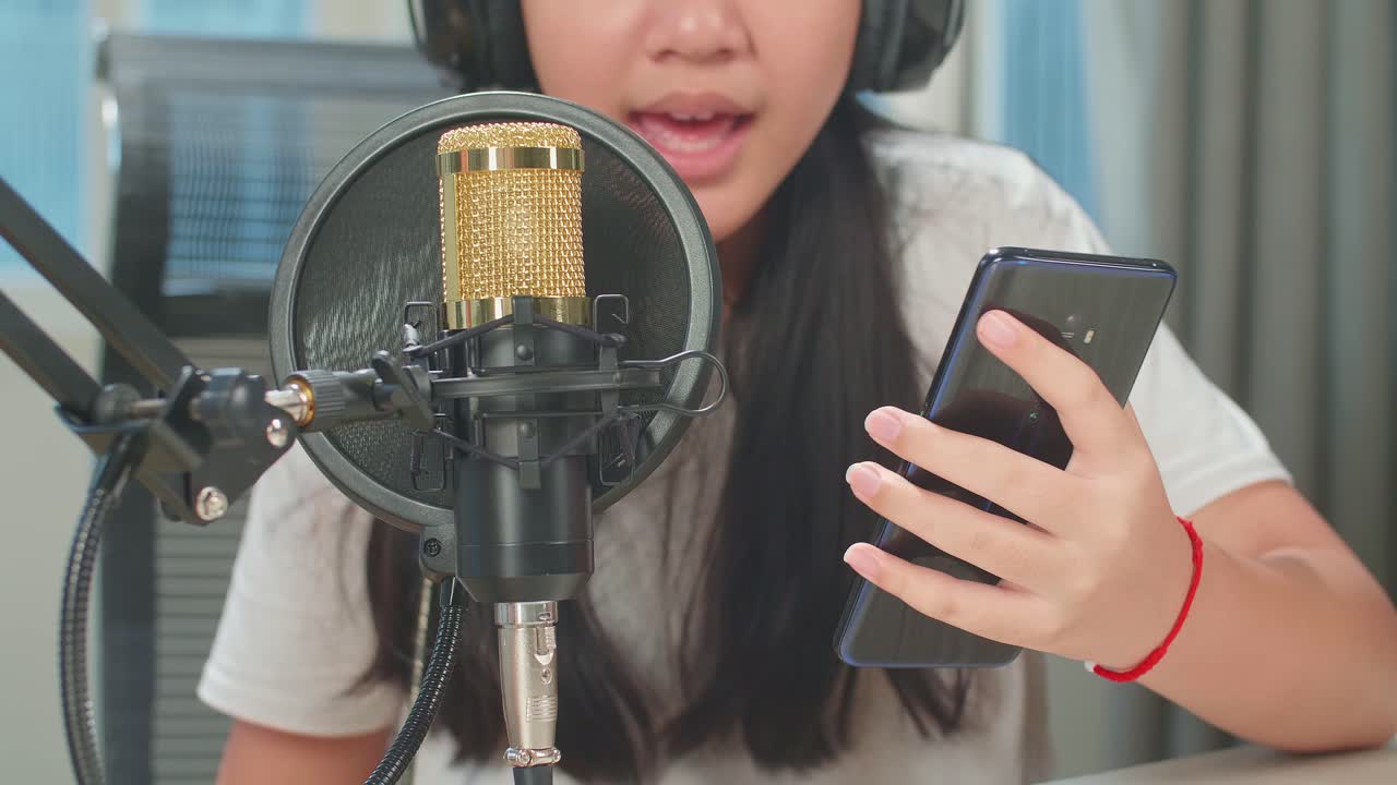 Asian Kid Girl Holding Mobile Phone And Singing With Microphone While Online Livestream In Podcast Studio