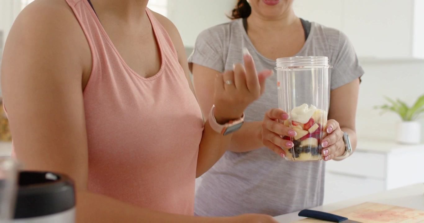 Dois adultos em roupas esportivas preparando um smoothie na bancada de casa com copo de liquidificador e faca.