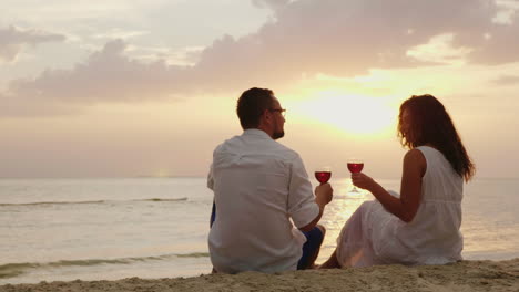 a young couple is drinking wine by the sea at sunset they sit on the sand clink glasses anniversary