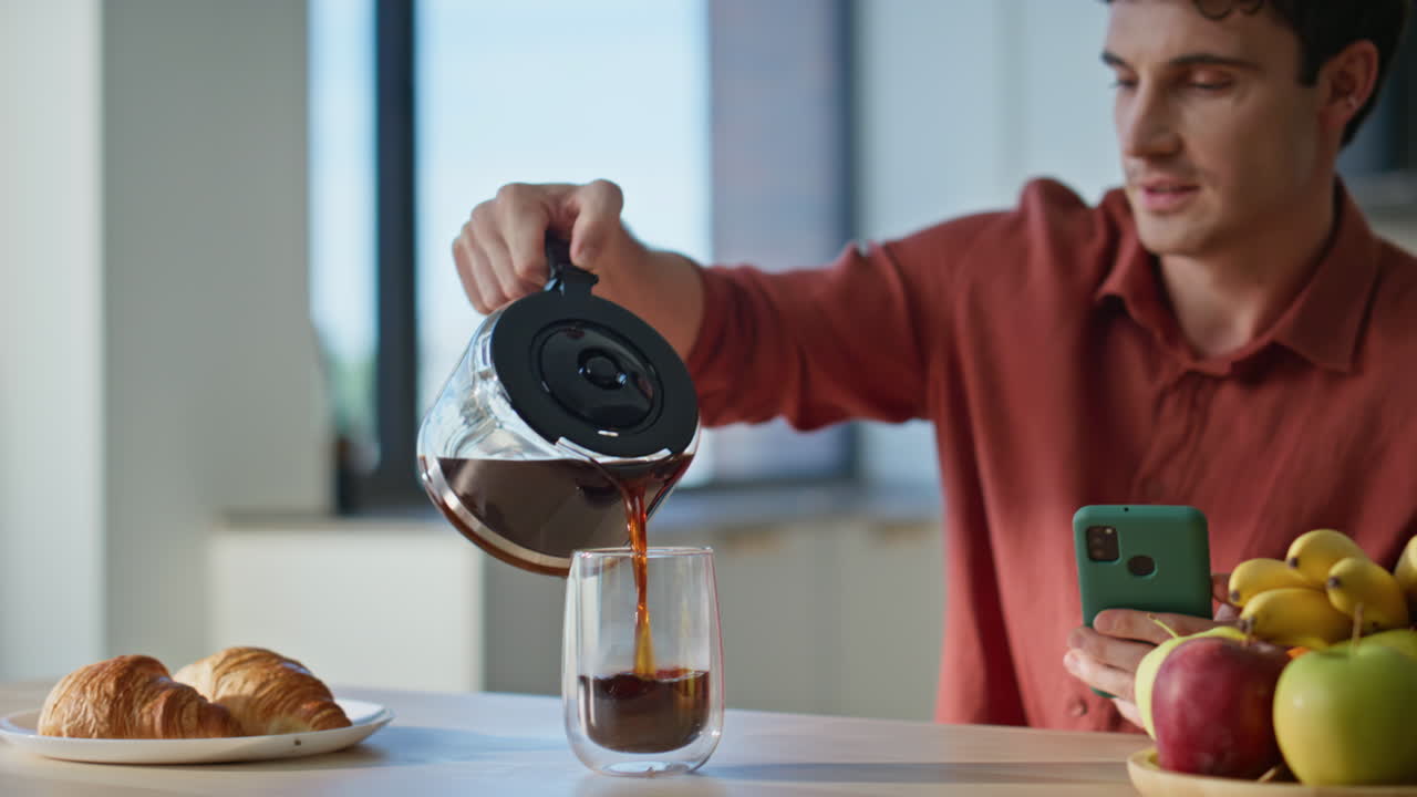 Relaxed man filling cup with brewed coffee holding smartphone at home closeup