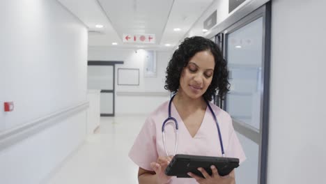 Focused-biracial-female-doctor-walking-and-using-tablet-in-hospital-in-slow-motion