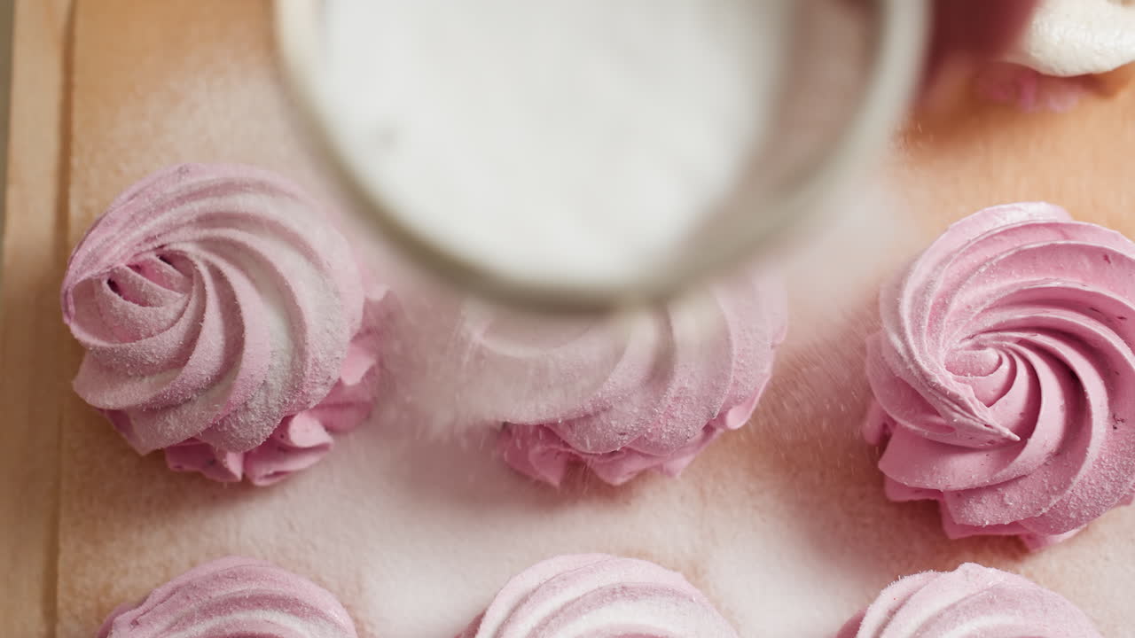Overhead view of prep process as powdered sugar is delicately sprinkled over swirl pink cupcakes on parchment paper, highlighting dessert creamy texture and sugary coating
