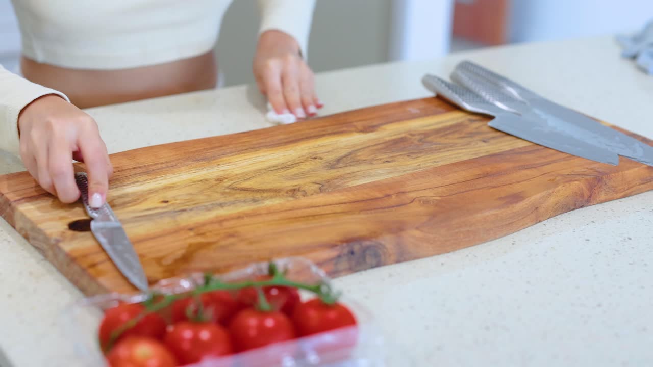 A person wipes a wooden cutting board with a cloth in a well-lit kitchen, surrounded by fresh tomatoes and knives