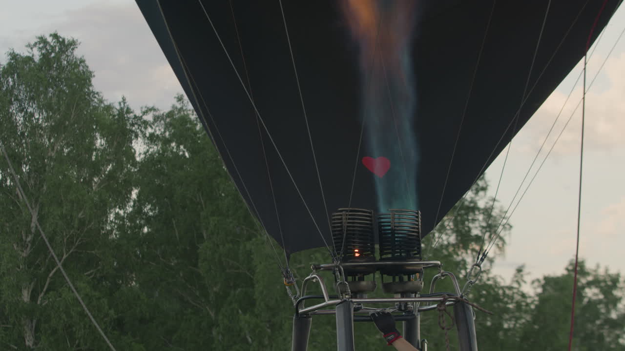 Powerful flame blasts upward into large colorful fabric envelope of hot air balloon during early inflation stage as burner fires with intensity, heart shape visible on lower gear, trees in background