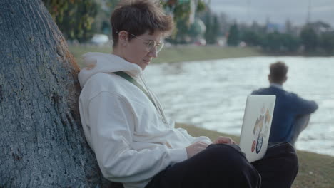 young woman sitting by tree outdoors using laptop