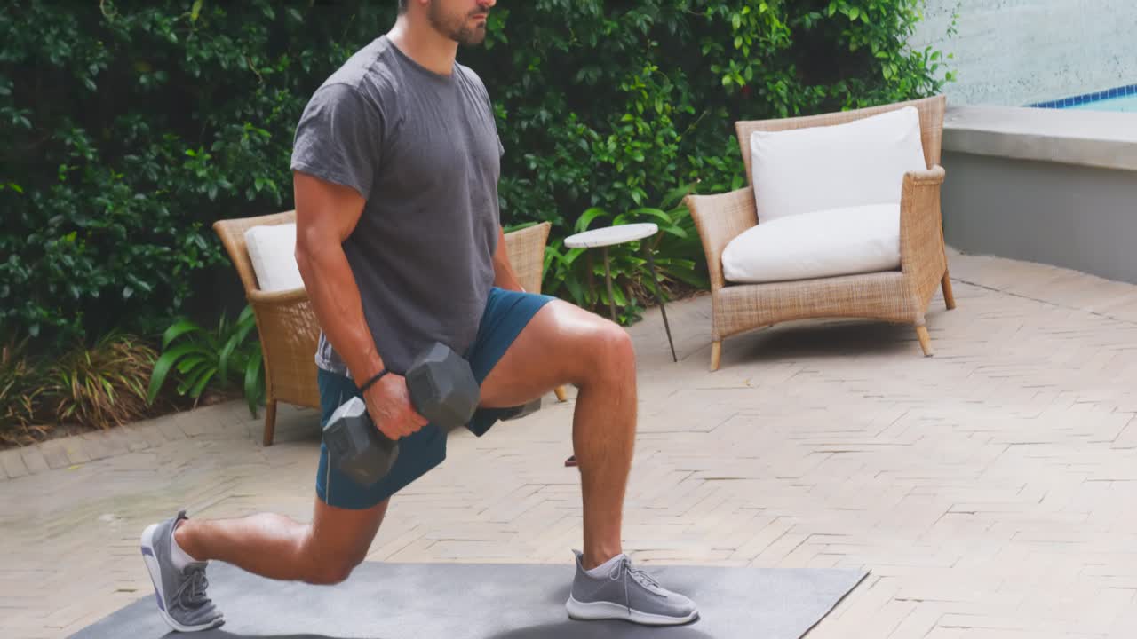 Erwachsener Mann tritt auf die Matte, macht Ausfallschritte mit Hanteln und trägt ein graues T-Shirt zum Krafttraining