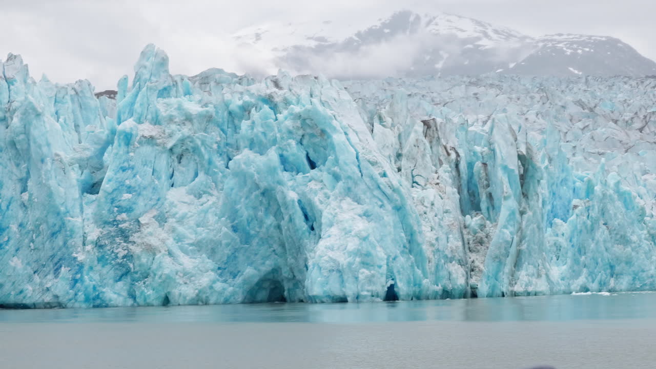 Panning shot from right to left of the Dawes Glacier in Endicott Arm in Alaska from a boat. It shows the mountains on the side and in the distance. It is a deep blue with spires and seagulls for scale