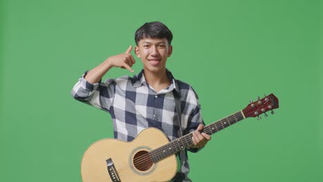 asian teenager with guitar showing call me gesture and smiling on green screen background