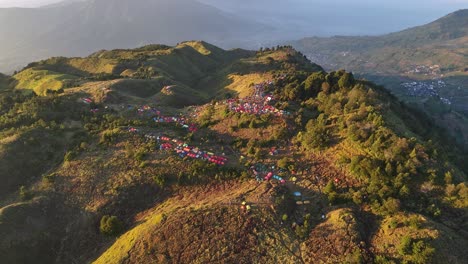 drone footage of a crowded mountaintop campsite at sunrise, featuring vibrant tents nestled among grassy peak and forested slopes — perfect for adventure and outdoor themes