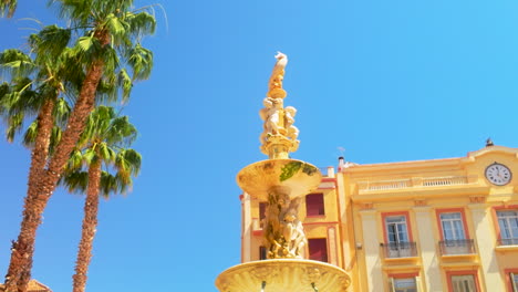 view of the fuente de génova in malaga, with its intricate statue and ornate design, framed by palm trees and historic buildings under a clear blue sky