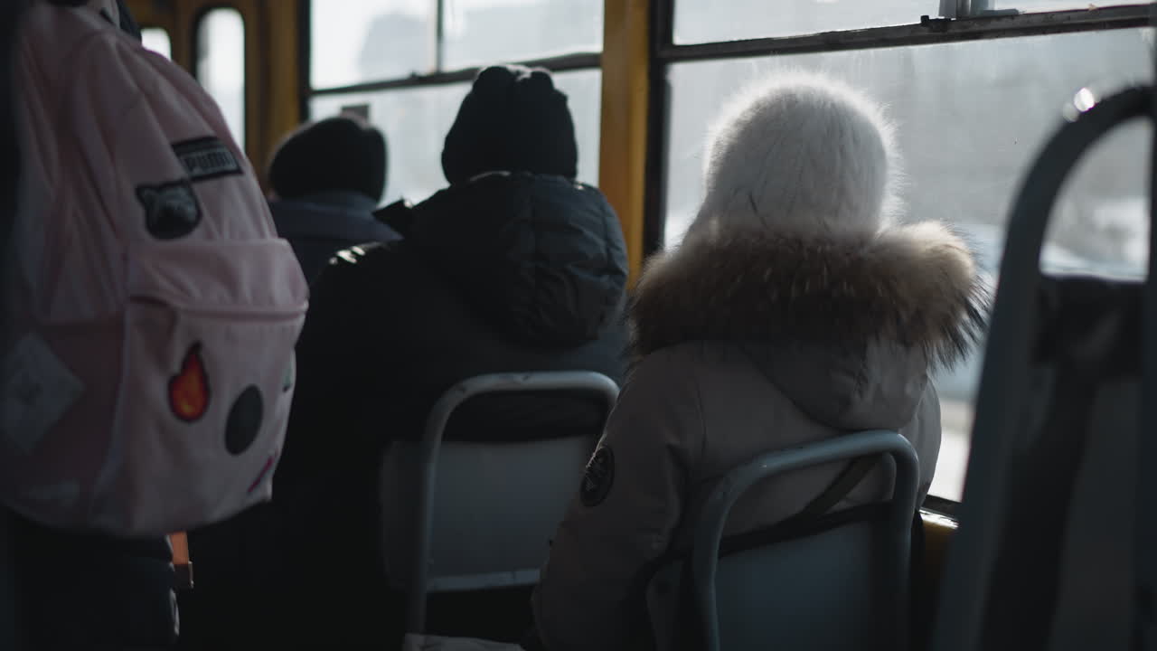 Back view of passengers in winter coats and beanies seated inside moving train, quiet commute with distant figures, bag on seat, subtle motion and chill across worn metal interior