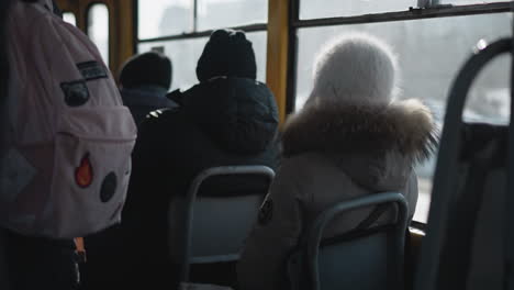 back view of passengers in winter coats and beanies seated inside moving train, quiet commute with distant figures, bag on seat, subtle motion and chill across worn metal interior