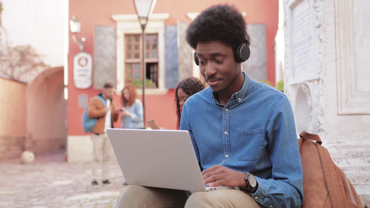 African american man making a video call with laptop while sitting on the ground in the street