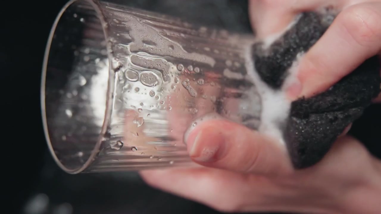 Primo piano di due mani che tengono un bicchiere da bibita trasparente a coste, mentre un panno scuro o una spugna strofina e risciacqua l'interno, lasciando schiuma di sapone, goccioline d'acqua, striature, riflessi e punti luce lucidi.