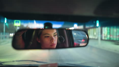 eine frau wird im rückspiegel ihres autos gesehen, während sie im verkehr wartet, mit der reflexion eines anderen autos, das hinter ihr durch den rückspiegel sichtbar ist, und verschwommenen stadtlichtern im hintergrund