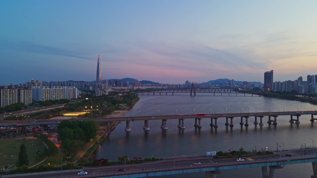 Un sorvolo aereo sopra il Parco Gwangnaru Hangang e il fiume Han dopo il tramonto rivela il Ponte Gwangjin e il Ponte Cheonho, con la Lotte World Tower che risplende nello skyline di Seul, Corea del Sud.