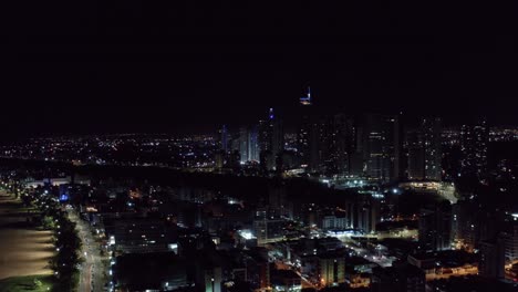 Right-trucking-aerial-night-shot-of-the-beautiful-tropical-beach-capital-cityscape-of-Joao-Pessoa,-Paraiba,-Brazil-from-the-Tambaú-neighborhood-with-a-beach-boardwalk,-busy-streets,-and-buildings