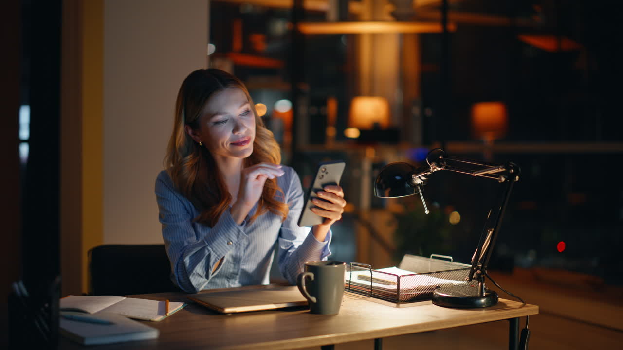 Lavoratore notturno che invia messaggi al cellulare seduto in un ufficio buio alla luce di una lampada, primo piano