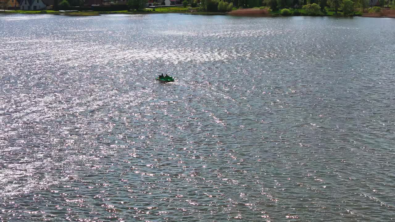 Epansive, high-angle wide-shot captures a lone dark boat on the shimmering, sun-dappled surface of Biržai's lake during a bright, sunny summer day, with distant green shores