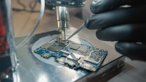 vista de cerca de las manos con guantes de un técnico usando un microscopio y pinzas de precisión para reparar una placa de circuitos detallada