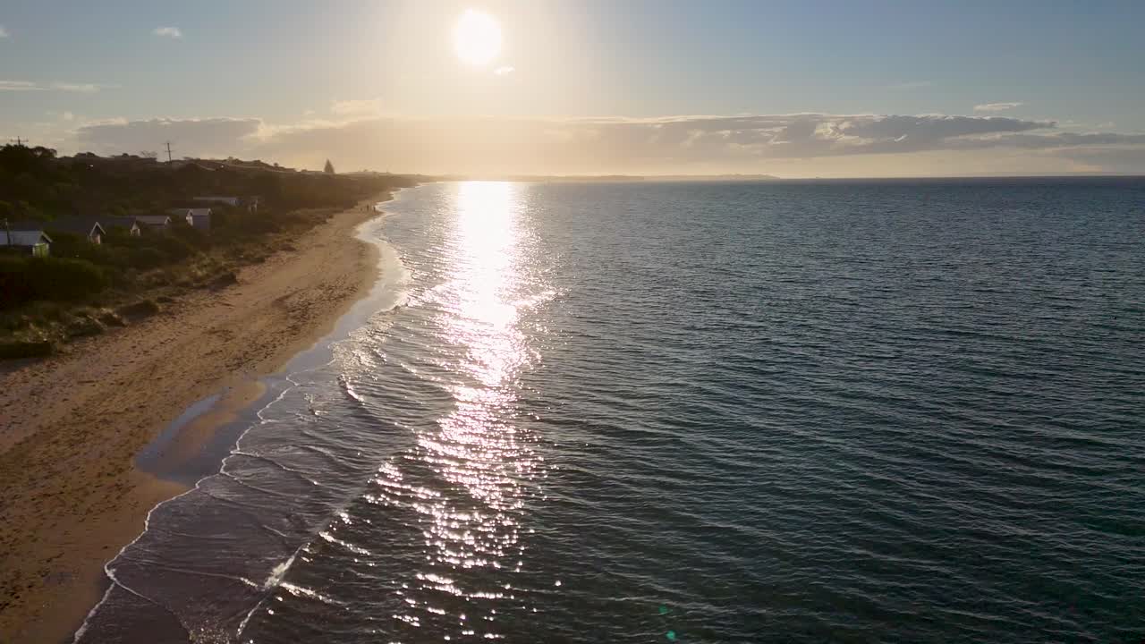 Drone sobrevoa a costa de areia, as cabines de praia e o mar calmo ao nascer do sol, com reflexos dourados