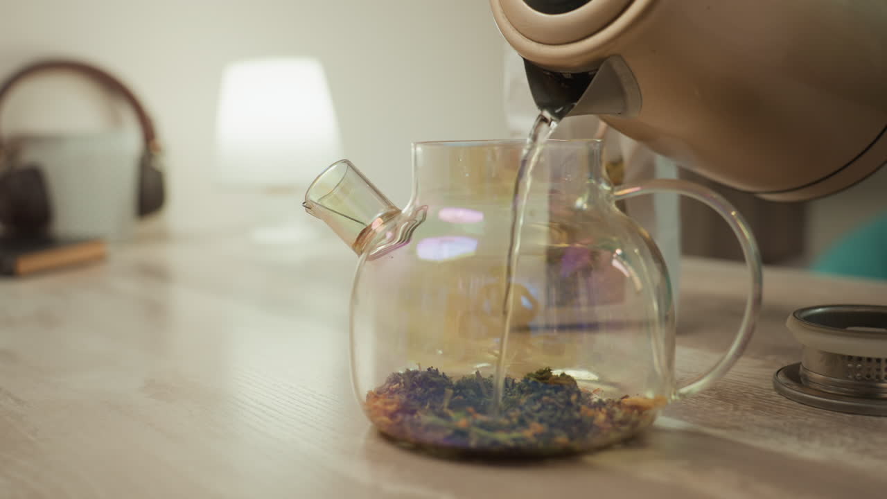 Teiera in vetro su tavolo di legno con tisana in infusione, una mano che versa dal bollitore, petali di fiori sfusi che si depositano, bagliore caldo di una lampada, cuffie e un ripiano accogliente, una scena intima di cura di sé che suggerisce un post-trattamento.