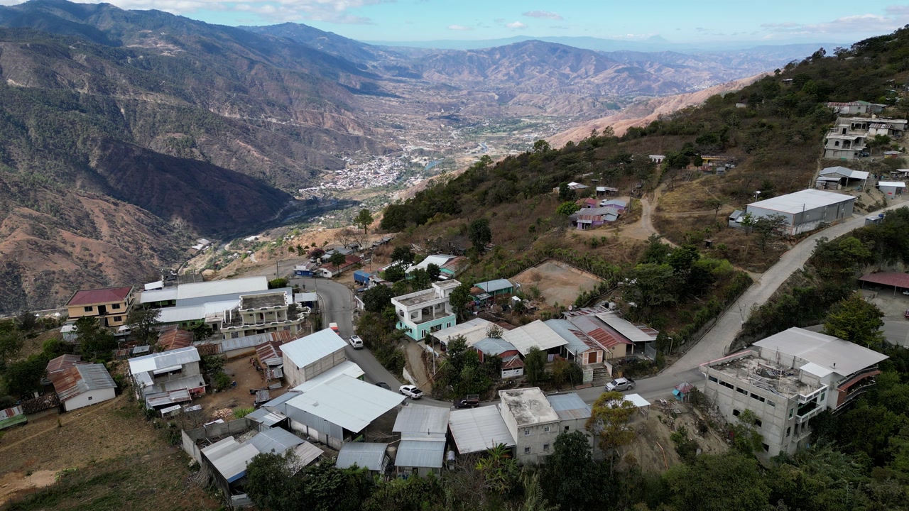 High-angle static drone shot of a hillside village near Sacapulas, El Quiche, Guatemala, highlighting the main road, residential clusters and layered mountain in the background.