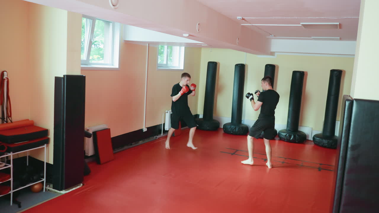 Two karate practitioners sparring in martial arts gym as one delivers strong front kick toward opponent who defends with gloves on red mat floor surrounded by punching bags during combat sport training