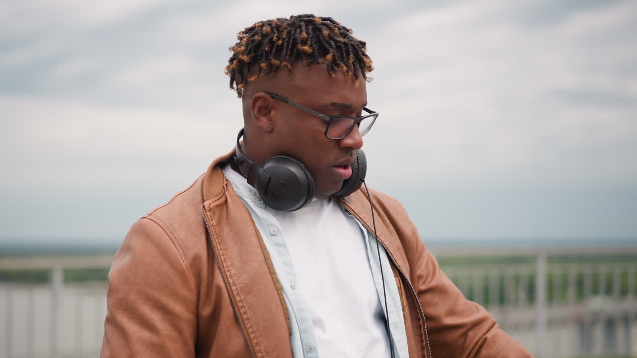 Sort DJ på tagterrasse med bærbar computer, fokuseret og sjælfuld, bærer hovedtelefoner og briller, læderjakke, farverig laptopkunst, overskyet himmel og fjern bysilhuet, intime nærbilleder der viser koncentration.