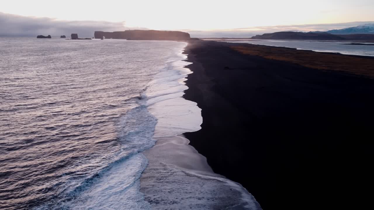 A tranquil black sand beach at Reynisfjara, Iceland, stretches into the distance, reflecting the soft hues of a sunset or sunrise over the calm North Atlantic waters