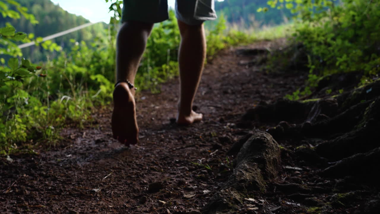 Shot of mans feet walking barefoot in slow motion on a path out of a dark forrest towards a bright green meadow in with sunshine