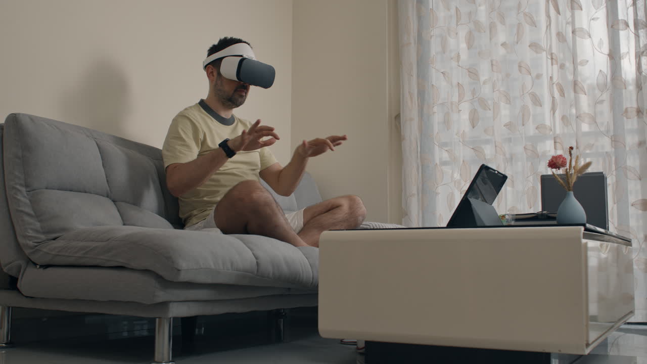 Man simulates playing piano or DJ set in air using VR headset while seated on couch at home